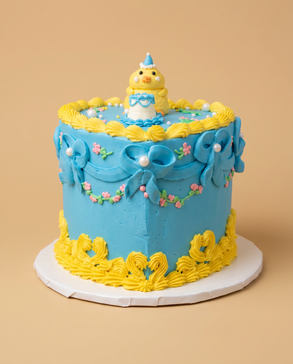Blue vintage buttercream cake with yellow piped shells, blue bows, pink floral garlands, and a fondant baby chick topper