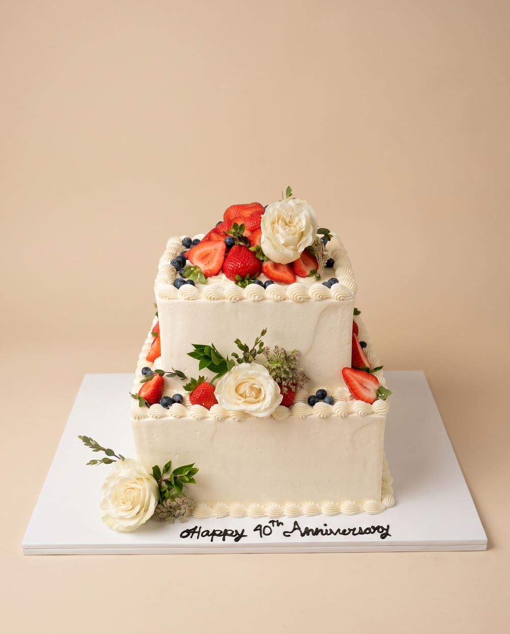 Two-tier square white buttercream anniversary cake topped with fresh strawberries, blueberries, and white roses
