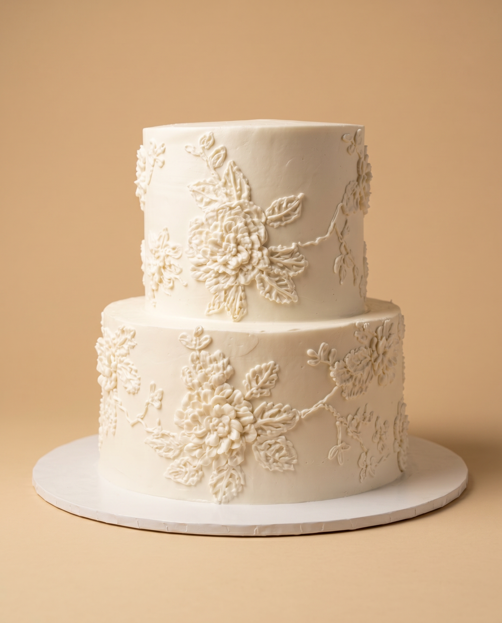 Two-tier ivory wedding cake with intricately piped buttercream flowers and leaves in matching tone-on-tone detail