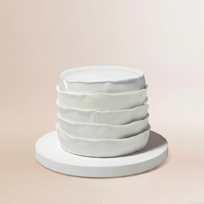 Single-tier round cake covered entirely in pure white whipped cream frosting, with five distinct horizontal bands of soft ruffled texture stacked from base to top, creating a sculptural tiered silhouette against a warm cream background