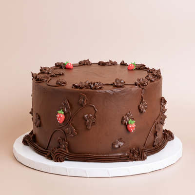 Round chocolate cake covered in smooth chocolate buttercream, decorated with piped chocolate leaves and vines, scalloped frosting borders, and small red fondant strawberries, on a white board against a beige background.
