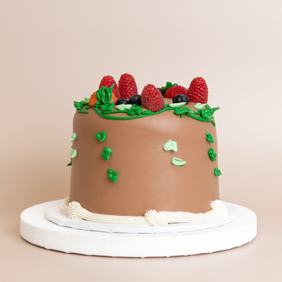 Round single-tier cake with milk chocolate whipped cream frosting, topped with fresh raspberries, blueberries, strawberries, and green buttercream leaves, finished with a white whipped cream base border