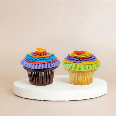 Two cupcakes — one chocolate, one vanilla — each topped with stacked tiers of ruffled rainbow buttercream resembling vibrant layered paper flowers