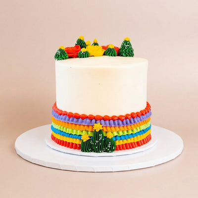 Single-tier white-frosted cake crowned with green buttercream cacti and red and yellow buttercream flowers, with rainbow serape-style ruffled bands in red, purple, orange, green, and blue wrapping the base