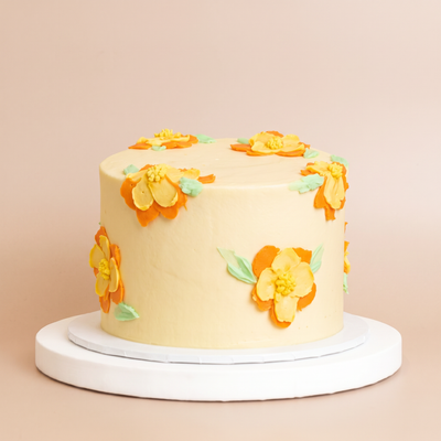 A round pale-yellow buttercream cake decorated with yellow and orange hand-piped flowers and green leaves, displayed on a white cake stand.