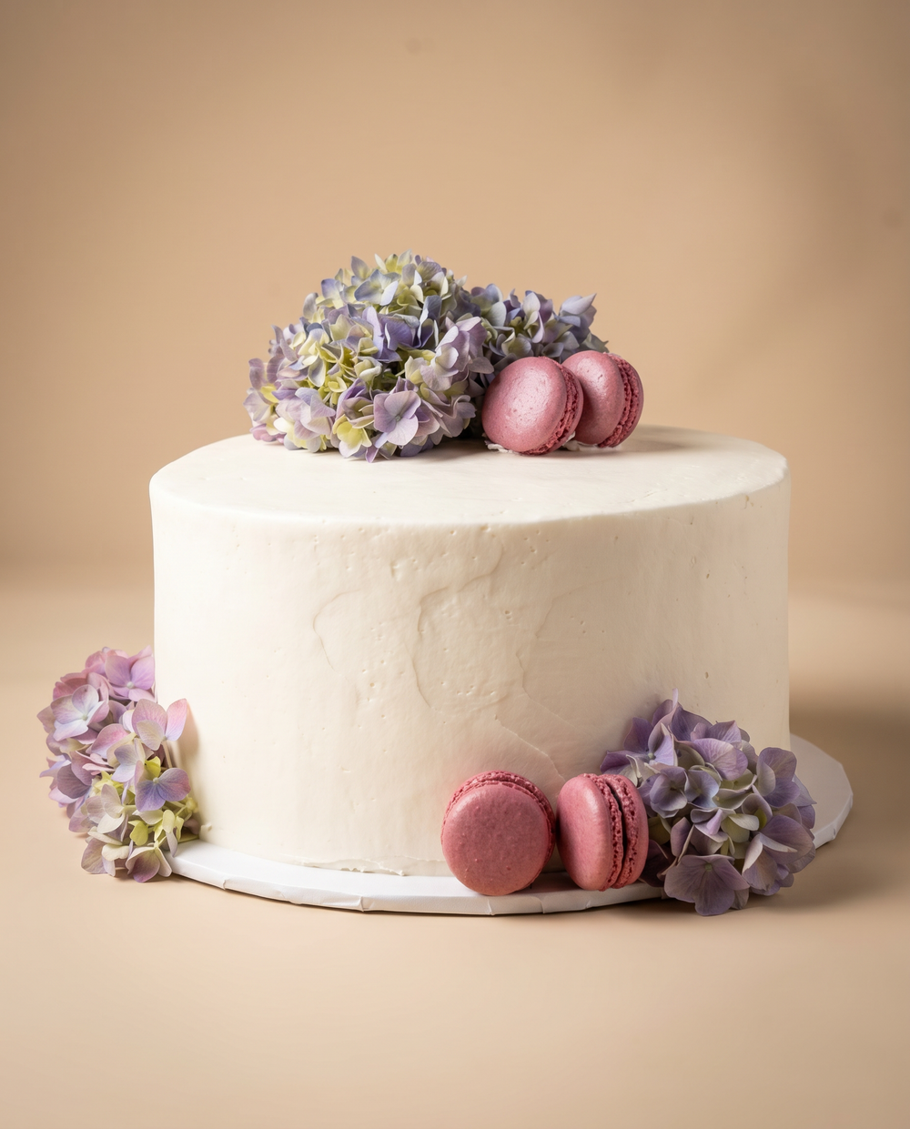 Single-tier white buttercream cake decorated with fresh purple hydrangeas and pink macarons