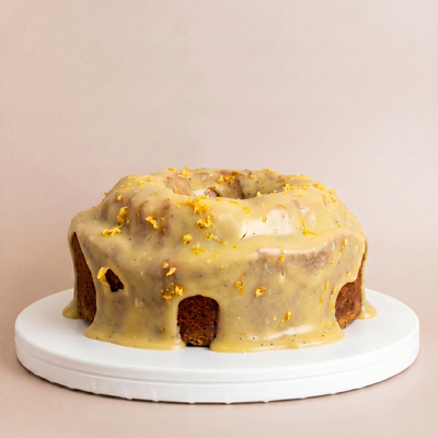 Whole round bundt cake on a white pedestal covered in thick glossy lemon poppy seed glaze with bright yellow lemon zest scattered on top