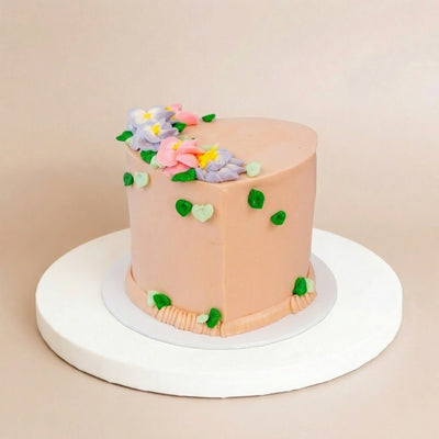 Tall heart-shaped cake with peachy-pink buttercream frosting, pastel buttercream flowers in pink, lavender, and yellow on top, and green leaf accents down the sides