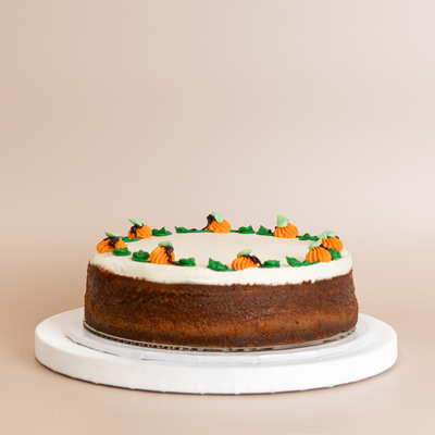 Short, wide cheesecake with golden-brown baked exterior and whipped cream top, decorated with piped orange buttercream pumpkins and green buttercream leaves