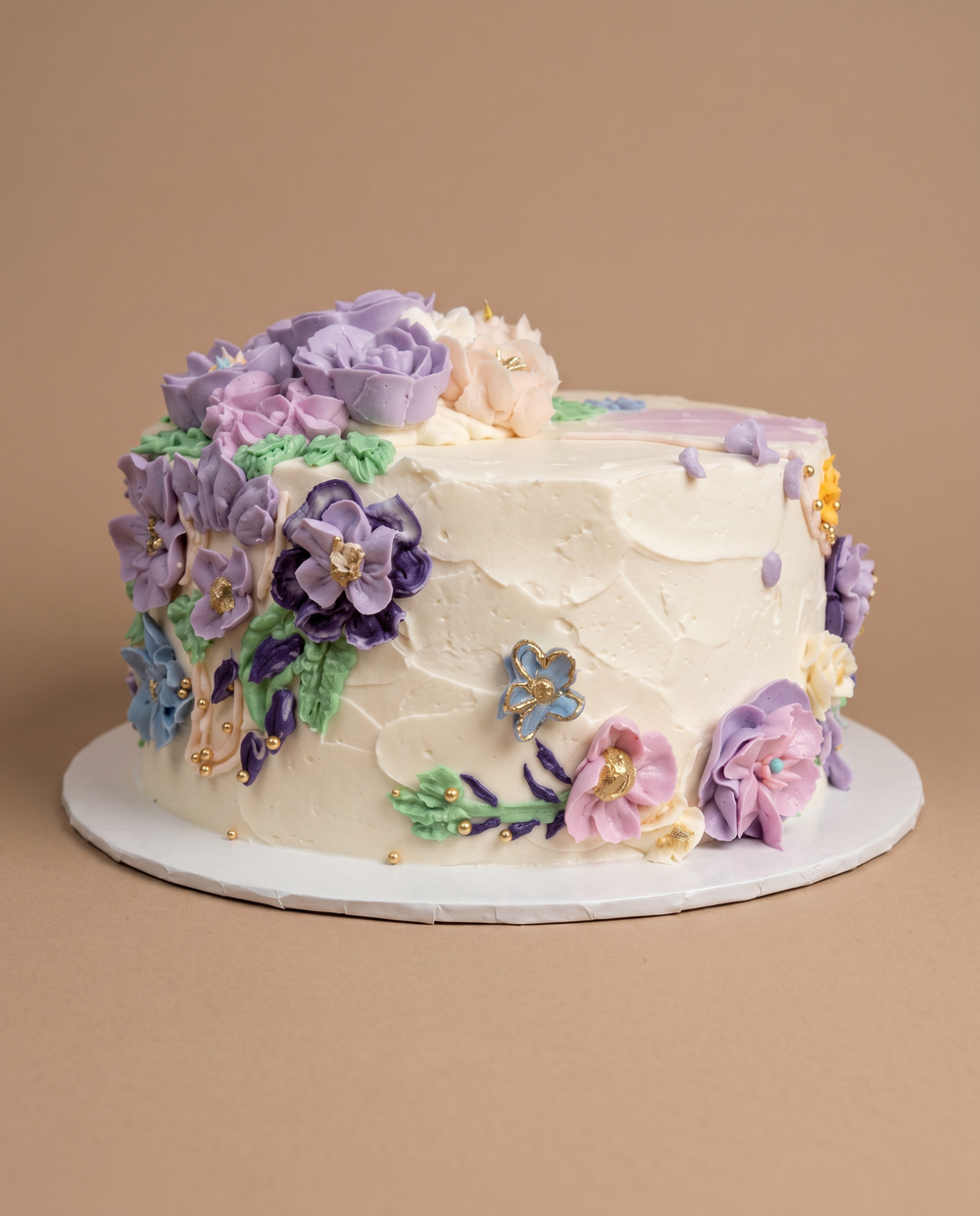White buttercream cake covered in piped purple and lavender flowers with gold pearl accents