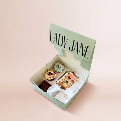 An open mint-green Sweet Lady Jane bakery box on a blush-pink background, with the lid propped up showing the 