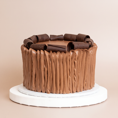Round chocolate cake with vertical hand-combed chocolate buttercream sides, topped with a ring of chocolate curls and shavings, on a white board against a beige background.