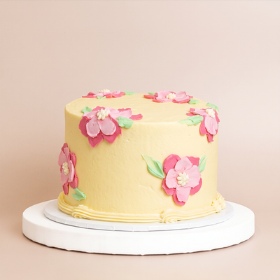 a round pale-yellow buttercream cake decorated with pink and magenta hand-piped flowers and green leaves, displayed on a white cake board.