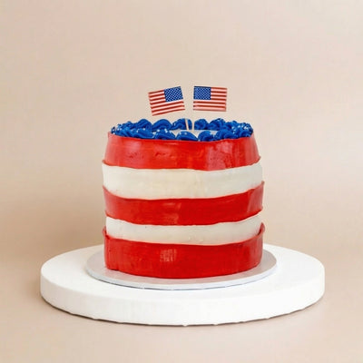 Tall round cake on a white pedestal with red and white stripe sides, blue rosette-piped top, star sprinkles, and two mini American flag toppers
