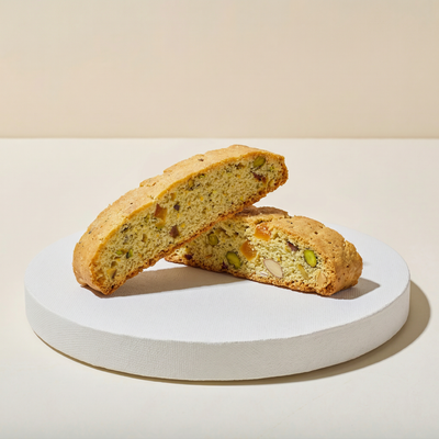 Two thick biscotti slices on a white pedestal with golden crisp interior studded with green pistachios and dried apricot