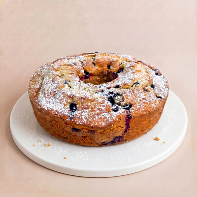 Whole round bundt cake on a white plate with golden-brown crust, dark blueberries peeking through, and a powdered sugar dusting