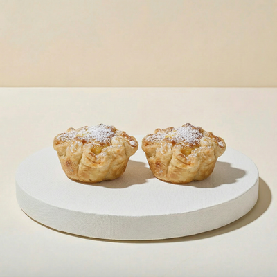 Two small golden puff pastries on a white pedestal with crisp flaky ruffled layers, cream cheese filling in the center, and powdered sugar