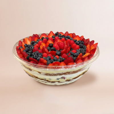Large clear bowl of layered trifle with cake, cream, and whipped cream visible through the sides, topped with an abundant mound of fresh mixed berries