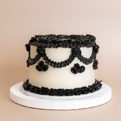 Round cake on a white pedestal with smooth ivory buttercream sides and ornate black piped rose, swag, and shell border decorations