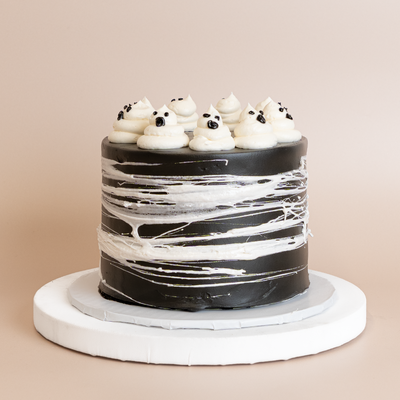 Tall round cake on a white pedestal with black buttercream sides, white spiderweb streaks, and piped white ghost figures with black dot eyes on top