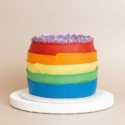 Tall round cake on a white pedestal with horizontal rainbow stripe buttercream in blue, green, yellow, orange, and red, and a purple rosette-piped top with rainbow sprinkles