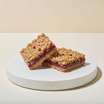 Two thick square oat bars on a white pedestal with golden oat streusel layers sandwiching vivid ruby-red raspberry jam filling