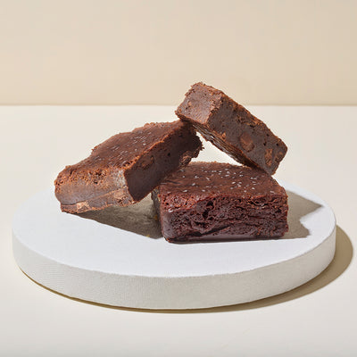 Three thick fudgy dark chocolate brownies stacked on a white pedestal, sprinkled with coarse sea salt